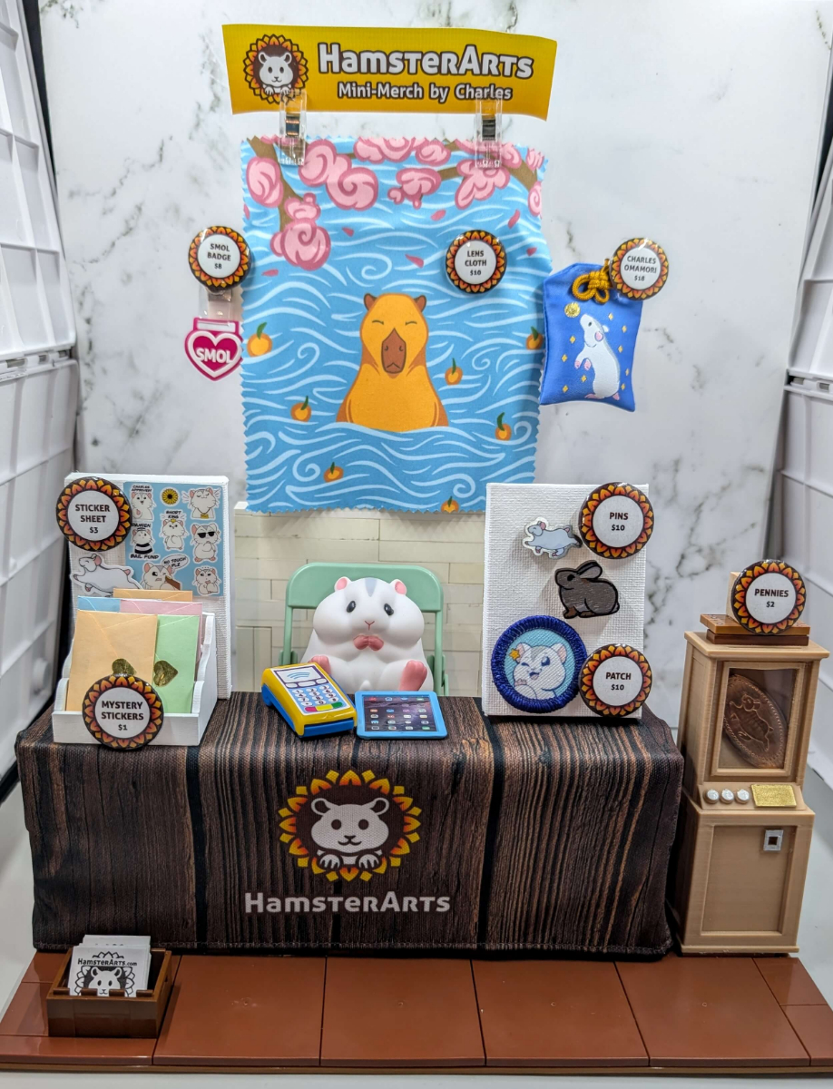 Charles the hamster (toy) sitting at a tiny artist booth, showing off some tiny pins and stickers he has for sale